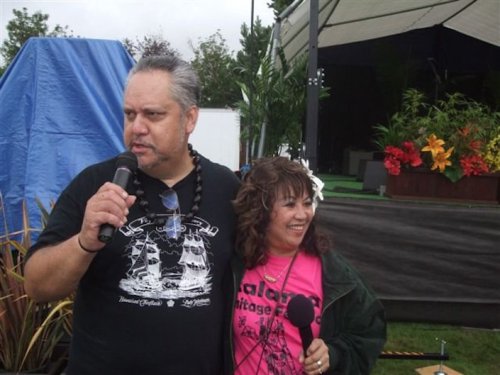 Two people stand together at an outdoor event, one holding a microphone, with a stage and flowers in the background.