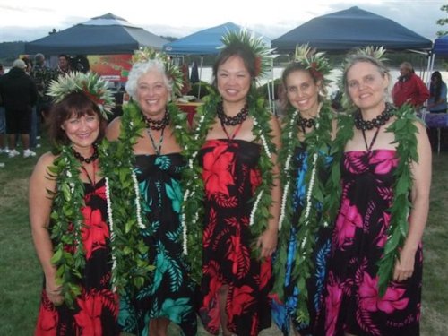 Five women in colorful floral dresses and leis pose together outdoors at a festive gathering.