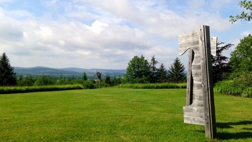A wooden sculpture stands on a grassy field, with rolling hills and trees in the background under a cloudy sky.