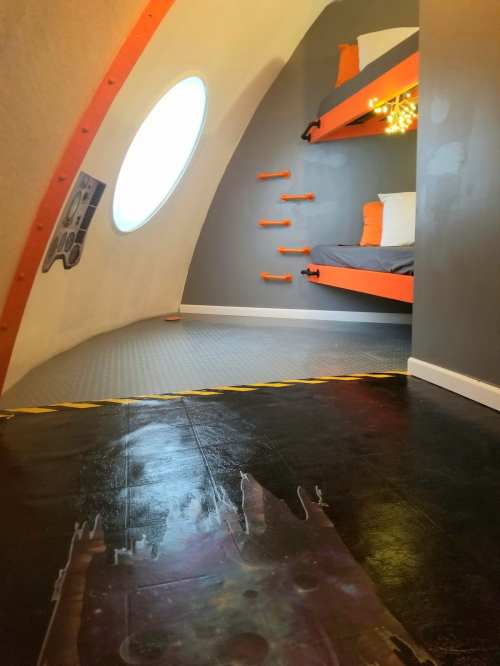 Interior of a futuristic room with curved walls, two orange bunk beds, and a round window, featuring a space-themed floor design.