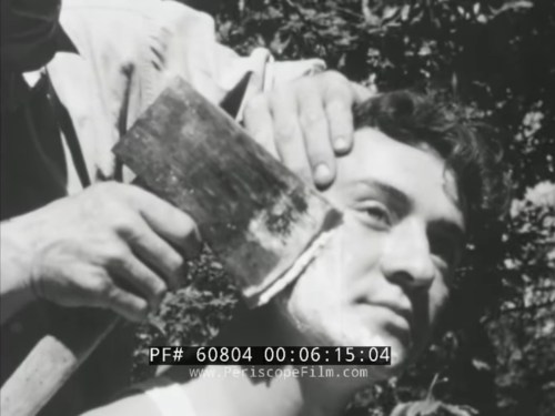A man is getting a traditional shave outdoors, with another man applying shaving cream and using a straight razor.