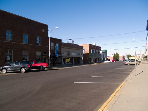 A quiet street scene featuring brick buildings, parked cars, and clear blue skies in a small town.