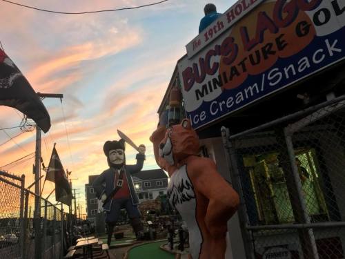 A colorful sunset over a miniature golf course featuring a pirate statue and a bear holding a drink.