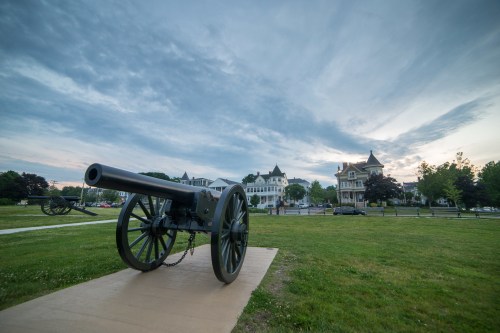 A historic cannon on a grassy field, with elegant buildings and a cloudy sky in the background.