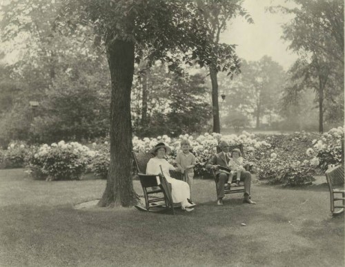 A vintage black-and-white photo of a woman and two children sitting on rocking chairs in a garden with blooming flowers.