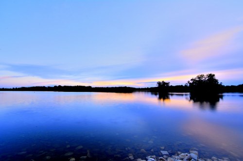 A serene lake at sunset, reflecting soft colors in the water with silhouettes of trees on the horizon.