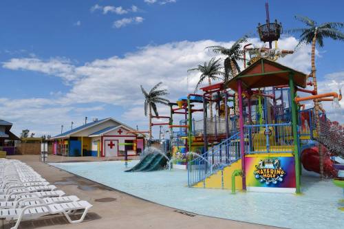 Colorful water park with slides, splash features, and lounge chairs under a blue sky with palm trees.