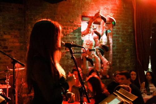 A singer performs on stage in a dimly lit venue, with colorful artwork and an audience in the background.