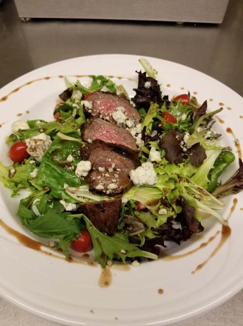 A plate of salad topped with sliced steak, cherry tomatoes, and blue cheese, drizzled with balsamic dressing.