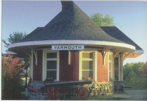 Historic round train station in Yarmouth, featuring a red exterior and stone foundation, surrounded by greenery.