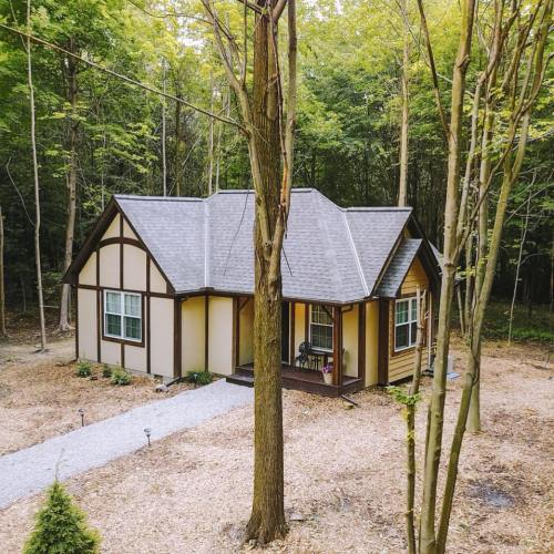 A cozy cottage nestled in a wooded area, surrounded by trees and a gravel pathway leading to the entrance.