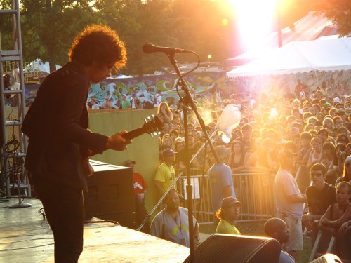 A musician plays guitar on stage as a crowd enjoys the performance, with sunlight creating a warm glow in the background.