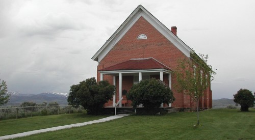 A brick building with a red roof, surrounded by green grass and trees, set against a cloudy sky and distant mountains.