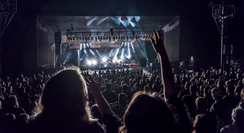 A large crowd at a concert with raised hands, illuminated by bright stage lights in a nighttime setting.