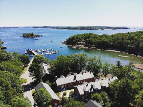 Aerial view of a serene coastal landscape with boats, lush greenery, and a calm blue bay under a clear sky.
