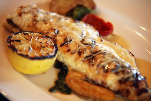 Grilled fish fillet topped with melted cheese, served with lemon, roasted potatoes, and garnished with herbs.