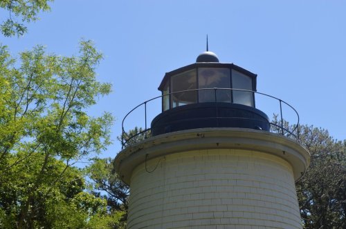 A close-up view of a lighthouse with a glass lantern room, surrounded by green trees and a clear blue sky.