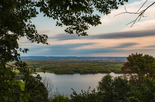 A serene landscape view of a river surrounded by lush greenery and distant hills under a colorful sky.