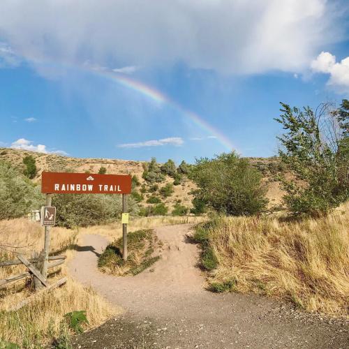 A sign for Rainbow Trail with a rainbow in the sky and a scenic landscape of hills and greenery in the background.
