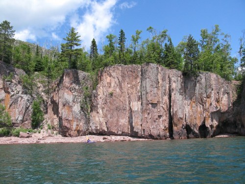 A rocky cliff rises above a calm lake, surrounded by lush green trees under a blue sky with scattered clouds.