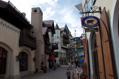 Charming village street with European-style architecture, shops, and a restaurant sign in a scenic outdoor setting.