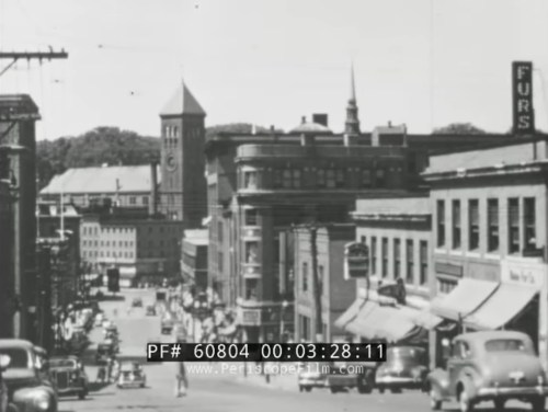 Black and white image of a busy street with vintage cars, shops, and a clock tower in the background.