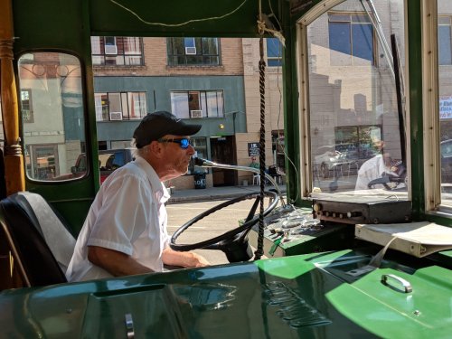 A bus driver in a cap sits at the wheel, looking out the front window of a vintage bus. Sunlight streams in.