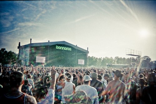 Crowd at a music festival with hands raised, facing a large stage under a bright sky.
