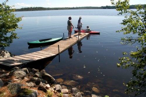 Three people by a wooden dock on a calm lake, with one in a kayak and two standing on the dock. Trees in the background.