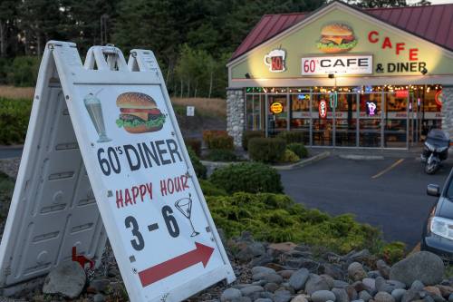 A sign for a 60's diner advertising happy hour, with a burger graphic, in front of a colorful diner building.