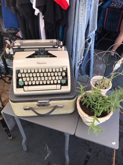 A vintage typewriter on a small table next to a potted plant, with clothing hanging in the background.