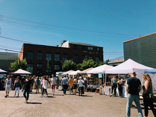 A bustling outdoor market with tents, vendors, and a crowd of people enjoying a sunny day.