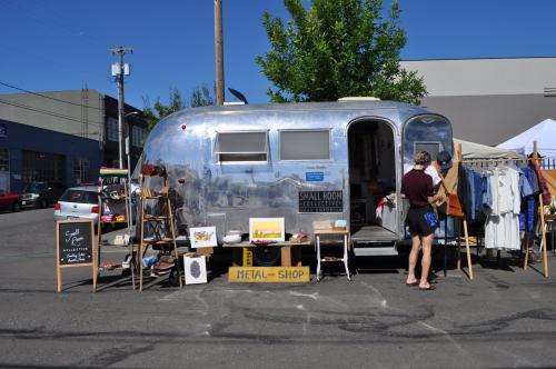 A shiny silver airstream trailer serves as a shop, with clothing and art displayed outside on a sunny day.