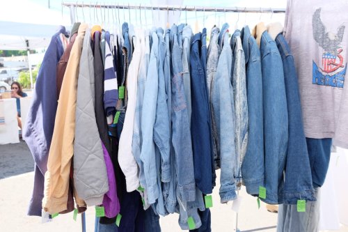 A rack of assorted jackets and denim hanging outdoors at a market, with colorful tags attached.