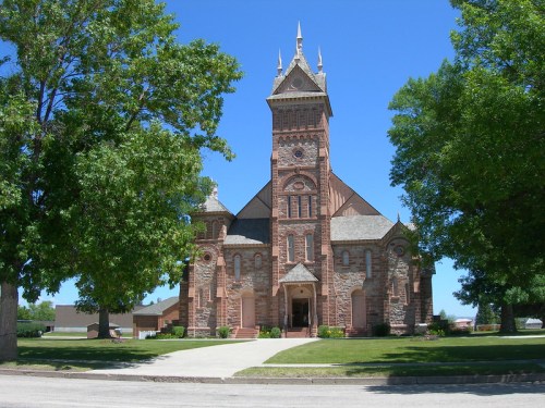 Historic stone church with a tall clock tower, surrounded by green trees and a clear blue sky.