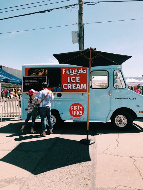 A blue ice cream truck with a sign reading "Fifty Licks" and people ordering ice cream on a sunny day.