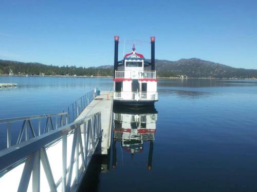 A paddleboat docked at a pier, reflecting in calm water with mountains in the background under a clear blue sky.