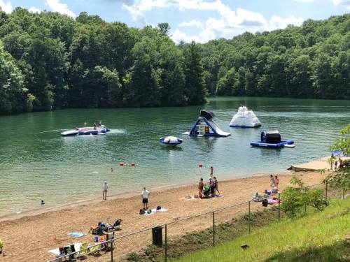 A sunny lakeside scene with inflatable water attractions, people swimming, and a sandy beach surrounded by trees.