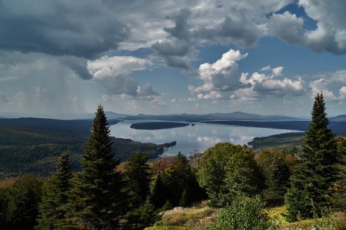 A scenic view of a lake surrounded by mountains and forests under a cloudy sky.