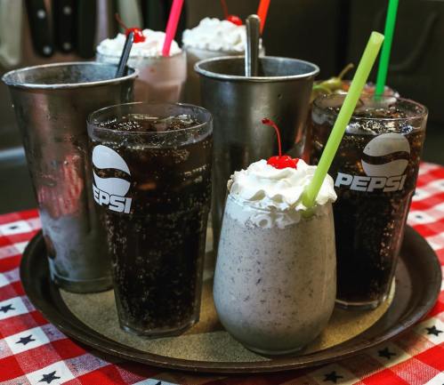 A tray with several glasses of soda and a milkshake topped with whipped cream and a cherry, set on a red and white checkered tablecloth.