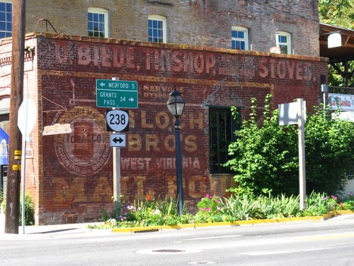 Faded vintage advertisement on a brick wall, with street signs for Grants Pass and Medford in a colorful setting.