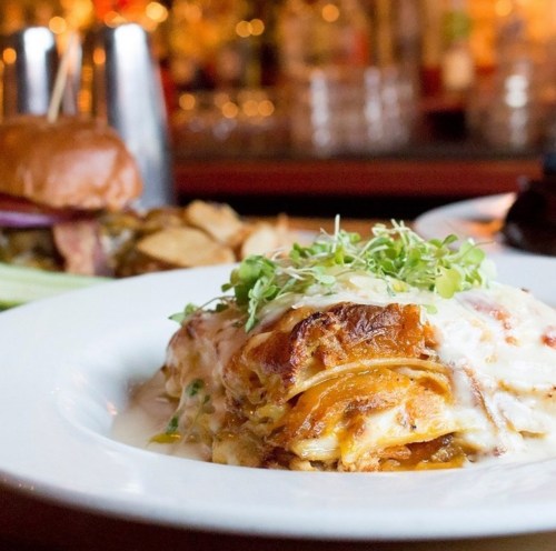 A plate of lasagna topped with fresh greens, served in a restaurant setting with a blurred background.