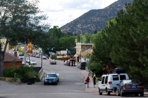 A small town street lined with trees, shops, and parked cars, set against a backdrop of mountains and cloudy skies.