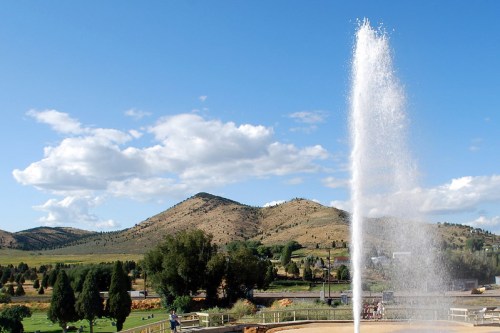 A water fountain sprays high into the air, with mountains and a blue sky in the background.
