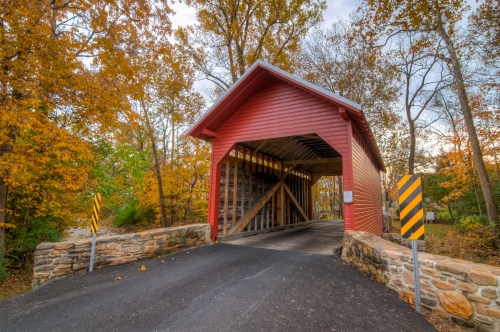 A red covered bridge surrounded by autumn trees with orange and yellow leaves, leading to a paved road.