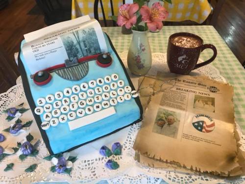 A vintage typewriter cake beside a coffee mug, flowers, and a newspaper with a "Breaking News" headline on a lace tablecloth.