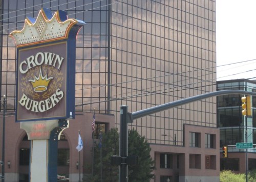 Sign for Crown Burgers in front of a modern glass building, with traffic lights and power lines in the foreground.