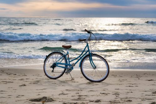 A blue bicycle stands on the sandy beach with waves gently crashing in the background under a cloudy sky.
