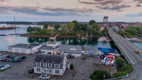 Aerial view of a coastal town with boats, a bridge, and a mix of buildings near the water under a cloudy sky.