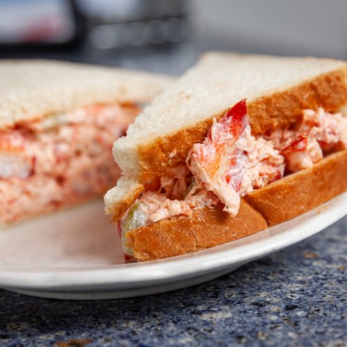 A close-up of a lobster salad sandwich on white bread, served on a plate with a blue countertop in the background.